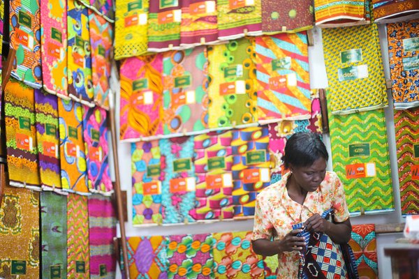 A trader measures a piece of GTP (Ghana Textile Print) cloth, by the yard, in her shop at Makola (mah-core-lah) Market, Accra. Photo by Yenkassa. CC BY.